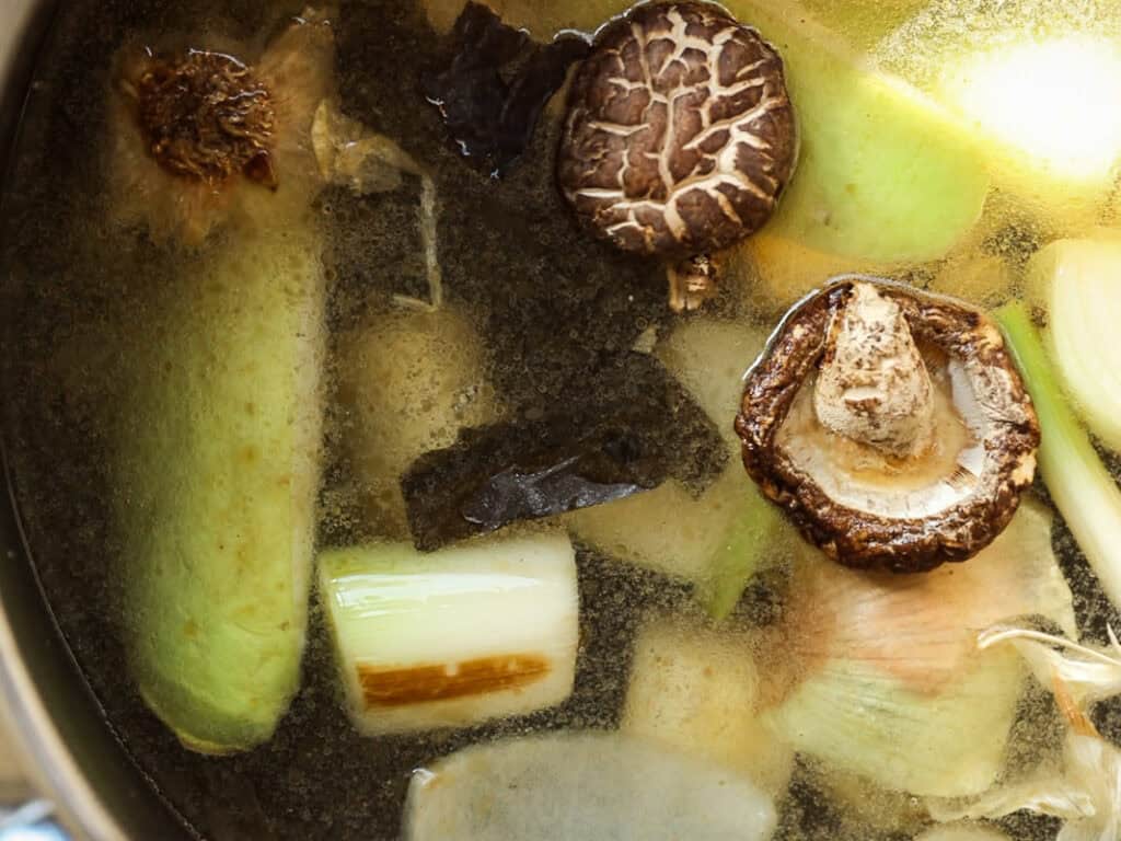 A pot filled with a variety of vegetables and mushrooms, including shiitake mushrooms, leeks, and seaweed, submerged in a clear broth. The ingredients are partially cooked, visible beneath the surface of the liquid.